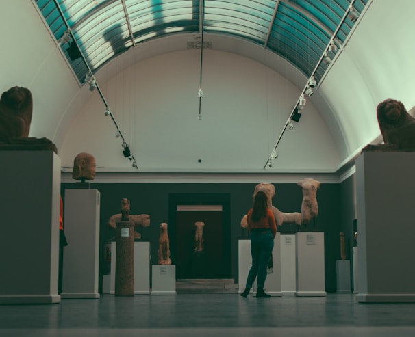 A person stands in an art gallery or museum hall observing sculptures that are displayed on pedestals. The sculptures appear to be ancient torso statues and heads made from stone. The hall has a high arched ceiling with skylights, and the lighting is subdued.