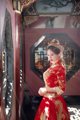 A woman wearing a traditional, intricately embroidered red and gold dress stands in an ornately decorated room. Sunlight streams through a window, illuminating her figure and creating dramatic lighting. The room features elegant woodwork and traditional architectural details, including geometric window designs and artistic ceiling elements.