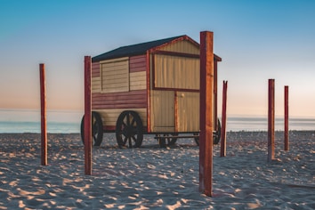 A wooden cart-like structure with black wheels is situated on a sandy beach. The cart is painted in shades of light yellow and red, and the roof is dark. Several wooden poles surround the cart, and the ocean is visible in the background under a clear blue sky.