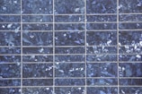 A close-up view of a solar panel composed of multiple square cells arranged in a grid pattern. The surface features a crystalline texture with a reflective quality, displaying various shades of blue and silver.
