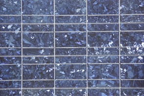 A close-up view of a solar panel composed of multiple square cells arranged in a grid pattern. The surface features a crystalline texture with a reflective quality, displaying various shades of blue and silver.