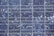 A close-up view of a solar panel composed of multiple square cells arranged in a grid pattern. The surface features a crystalline texture with a reflective quality, displaying various shades of blue and silver.