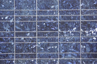 A close-up view of a solar panel composed of multiple square cells arranged in a grid pattern. The surface features a crystalline texture with a reflective quality, displaying various shades of blue and silver.