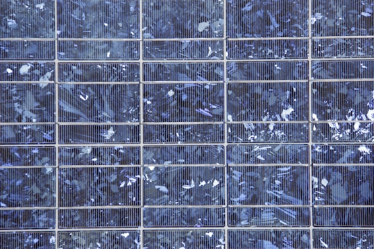 A close-up view of a solar panel composed of multiple square cells arranged in a grid pattern. The surface features a crystalline texture with a reflective quality, displaying various shades of blue and silver.