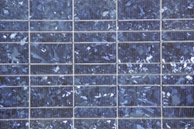 A close-up view of a solar panel composed of multiple square cells arranged in a grid pattern. The surface features a crystalline texture with a reflective quality, displaying various shades of blue and silver.