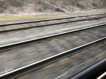 Multiple railway tracks extend into the distance. The tracks are captured from a moving perspective, creating a sense of motion. The surrounding area features earthy tones with sparse vegetation on either side.