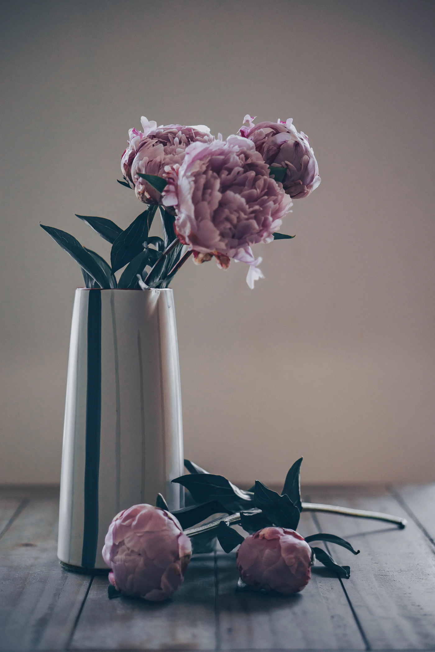 Pale peonies in a tall ceramic vessel, with two stems resting on the table