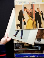 A stack of vintage vinyl record covers is held by a hand against a dark background. The top album is titled 's marvelous', featuring people dressed in formal attire posing playfully with poles. The scene captures a retro and lively atmosphere.