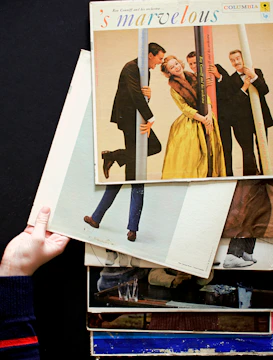 A stack of vintage vinyl record covers is held by a hand against a dark background. The top album is titled 's marvelous', featuring people dressed in formal attire posing playfully with poles. The scene captures a retro and lively atmosphere.
