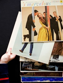 A stack of vintage vinyl record covers is held by a hand against a dark background. The top album is titled 's marvelous', featuring people dressed in formal attire posing playfully with poles. The scene captures a retro and lively atmosphere.