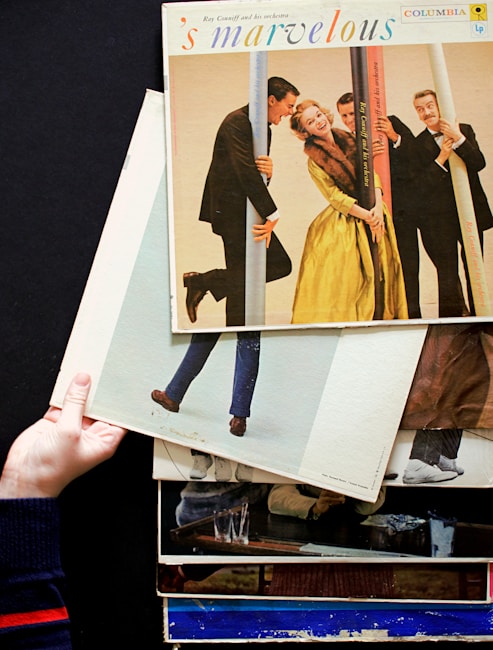 A stack of vintage vinyl record covers is held by a hand against a dark background. The top album is titled 's marvelous', featuring people dressed in formal attire posing playfully with poles. The scene captures a retro and lively atmosphere.