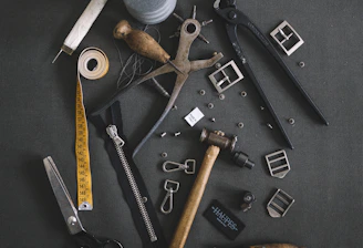 Various tailoring tools are scattered on a dark fabric background. Among these tools are scissors, a measuring tape, a spool of thread, a zipper, metal buckles, a wooden mallet, pliers, cutters, and other sewing accessories.