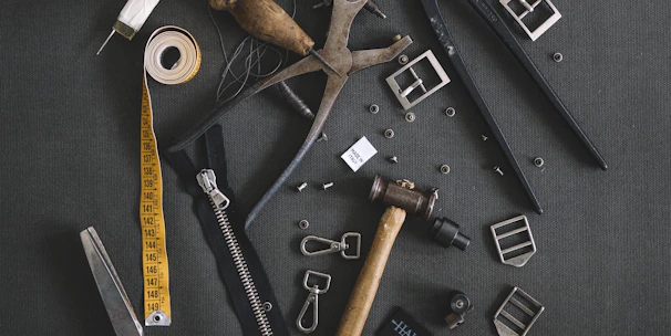 Various tailoring tools are scattered on a dark fabric background. Among these tools are scissors, a measuring tape, a spool of thread, a zipper, metal buckles, a wooden mallet, pliers, cutters, and other sewing accessories.