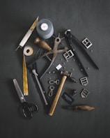 Various tailoring tools are scattered on a dark fabric background. Among these tools are scissors, a measuring tape, a spool of thread, a zipper, metal buckles, a wooden mallet, pliers, cutters, and other sewing accessories.