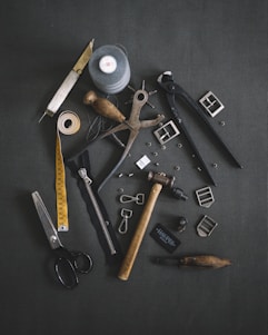 Various tailoring tools are scattered on a dark fabric background. Among these tools are scissors, a measuring tape, a spool of thread, a zipper, metal buckles, a wooden mallet, pliers, cutters, and other sewing accessories.