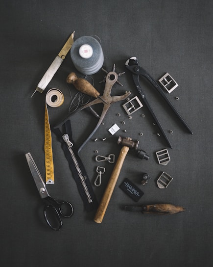 Various tailoring tools are scattered on a dark fabric background. Among these tools are scissors, a measuring tape, a spool of thread, a zipper, metal buckles, a wooden mallet, pliers, cutters, and other sewing accessories.