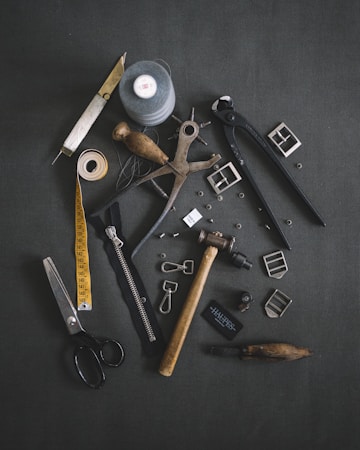 Various tailoring tools are scattered on a dark fabric background. Among these tools are scissors, a measuring tape, a spool of thread, a zipper, metal buckles, a wooden mallet, pliers, cutters, and other sewing accessories.