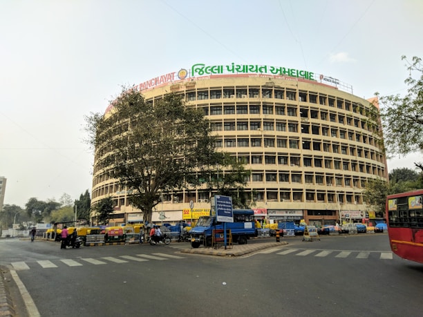 A large, circular multi-story building with numerous windows and a banner featuring text in multiple languages on top. In front of the building, there are several auto rickshaws, motorcycles, and people on the street, indicating a busy urban setting. There are trees on both sides of the building and a red public bus can be seen on the right.