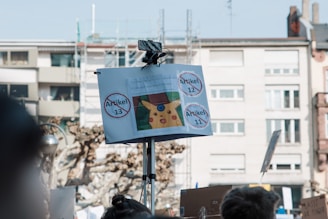 A protest scene featuring a sign mounted on a pole displaying images and the words 'Artikel 13,' 'Artikel 12,' and 'Artikel 11' inside circles with a strike through them. There is a cartoon character visible on the poster. The background shows a residential building and some bare trees.