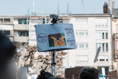 A protest scene featuring a sign mounted on a pole displaying images and the words 'Artikel 13,' 'Artikel 12,' and 'Artikel 11' inside circles with a strike through them. There is a cartoon character visible on the poster. The background shows a residential building and some bare trees.