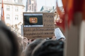 A person holds up a protest sign made of cardboard with a drawing of a video player icon featuring a sad face emoji. The text in German suggests the demonstration cannot be shown due to copyright reasons. The background includes the facade of historic buildings and blurry protest signs held by others, indicating a rally or demonstration environment.
