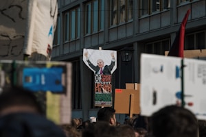 A crowded street scene with a protest in progress, featuring people holding signs and banners. A notable poster depicts a caricature of a person with raised arms, surrounded by words like 'fake news' and 'upload filter'. The background includes a building with multiple windows.