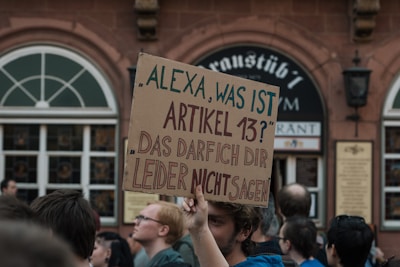 A man holds up a large cardboard sign in a crowd. The sign is written in German, asking 'Alexa, what is Article 13?' and stating 'Unfortunately, I cannot tell you that.' People around the man appear to be participating in a public demonstration. The background includes a building with arched windows and a sign indicating it is a restaurant.
