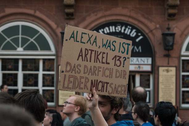A man holds up a large cardboard sign in a crowd. The sign is written in German, asking 'Alexa, what is Article 13?' and stating 'Unfortunately, I cannot tell you that.' People around the man appear to be participating in a public demonstration. The background includes a building with arched windows and a sign indicating it is a restaurant.