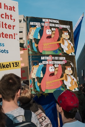 Protesters are gathered, holding signs with cartoons and text regarding Article 13 and Article 11. The visible sign includes a cartoon character holding a trumpet and a message in German related to internet regulations.