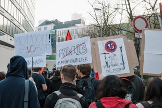 A group of people is participating in a protest, holding signs with messages against Article 13 and automated bots. One sign features binary code and a hashtag declaring 'NO BOTS', while another reads 'ARTIKEL 13' with 'NO UPLOAD FILTER'. A third sign displays a graphic with the text 'YOU SHALL NOT BE PASSED'. The crowd is gathered on a street lined with buildings and trees, and it appears to be daytime.