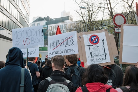 A group of people is participating in a protest, holding signs with messages against Article 13 and automated bots. One sign features binary code and a hashtag declaring 'NO BOTS', while another reads 'ARTIKEL 13' with 'NO UPLOAD FILTER'. A third sign displays a graphic with the text 'YOU SHALL NOT BE PASSED'. The crowd is gathered on a street lined with buildings and trees, and it appears to be daytime.