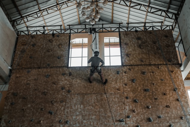 A person wearing a harness is climbing an indoor rock wall. The wall has various holds and is set in a building with a high ceiling, featuring large windows and a metal roof structure. A chandelier hangs from the ceiling, adding an interesting contrast to the sports setting.