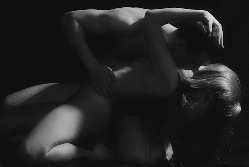 A black and white photograph capturing an intimate moment between two individuals. The lighting is dramatic, highlighting the contours and textures of their bodies. The scene is both intimate and artistic, focusing on the human form and emotions involved.