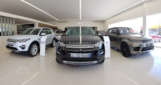 A sleek and modern car showroom featuring a line of luxury SUVs. The vehicles are displayed on a glossy white tiled floor under bright lighting, which enhances their metallic finishes. The cars are positioned in a spacious and well-lit environment, possibly indicating a high-end automobile dealership.