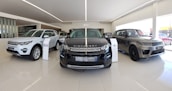 A sleek and modern car showroom featuring a line of luxury SUVs. The vehicles are displayed on a glossy white tiled floor under bright lighting, which enhances their metallic finishes. The cars are positioned in a spacious and well-lit environment, possibly indicating a high-end automobile dealership.
