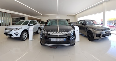 A sleek and modern car showroom featuring a line of luxury SUVs. The vehicles are displayed on a glossy white tiled floor under bright lighting, which enhances their metallic finishes. The cars are positioned in a spacious and well-lit environment, possibly indicating a high-end automobile dealership.