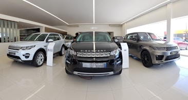 A sleek and modern car showroom featuring a line of luxury SUVs. The vehicles are displayed on a glossy white tiled floor under bright lighting, which enhances their metallic finishes. The cars are positioned in a spacious and well-lit environment, possibly indicating a high-end automobile dealership.