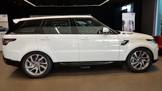A sleek white luxury SUV is parked indoors, possibly in a showroom. The vehicle has large alloy wheels and tinted windows. The environment suggests a premium and modern setting, with banners in the background hinting at branding or promotional material.