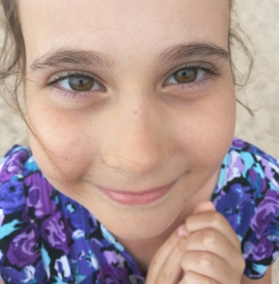 A close-up of a smiling child's face with expressive brown eyes and rosy cheeks, wearing a colorful floral-patterned shirt. The hands are clasped together in front of the chin, conveying a sense of innocence and charm.
