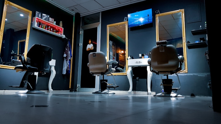 A modern barbershop interior with several styling chairs facing large ornate mirrors. The room is dimly lit, creating a calm ambiance. A television is mounted on the wall above the central mirror, and shelves with hair products are visible. A person stands in the doorway, possibly a barber or customer.