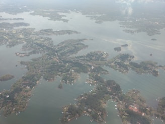 An aerial view of a large body of water with various winding coastlines densely populated with green foliage. Several small islands and peninsulas are evenly scattered throughout the water. Residential areas with numerous houses and docks are visible along the shorelines, and the overall landscape is surrounded by more expansive land areas that disappear into the horizon.