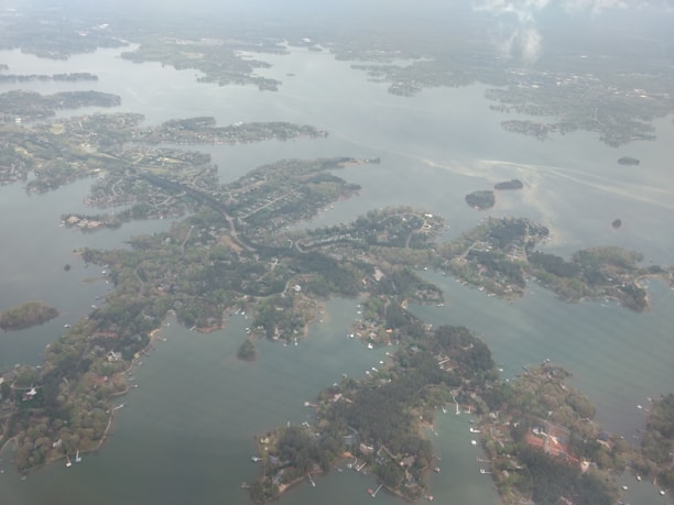 An aerial view of a large body of water with various winding coastlines densely populated with green foliage. Several small islands and peninsulas are evenly scattered throughout the water. Residential areas with numerous houses and docks are visible along the shorelines, and the overall landscape is surrounded by more expansive land areas that disappear into the horizon.