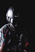 A person wearing a detailed motorcycle helmet and colorful gloves is set against a dark background. The helmet has intricate designs, and the gloves feature vibrant patterns. The lighting creates a dramatic, moody effect.