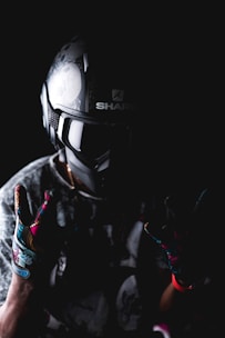 A person wearing a detailed motorcycle helmet and colorful gloves is set against a dark background. The helmet has intricate designs, and the gloves feature vibrant patterns. The lighting creates a dramatic, moody effect.