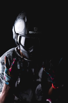 A person wearing a detailed motorcycle helmet and colorful gloves is set against a dark background. The helmet has intricate designs, and the gloves feature vibrant patterns. The lighting creates a dramatic, moody effect.