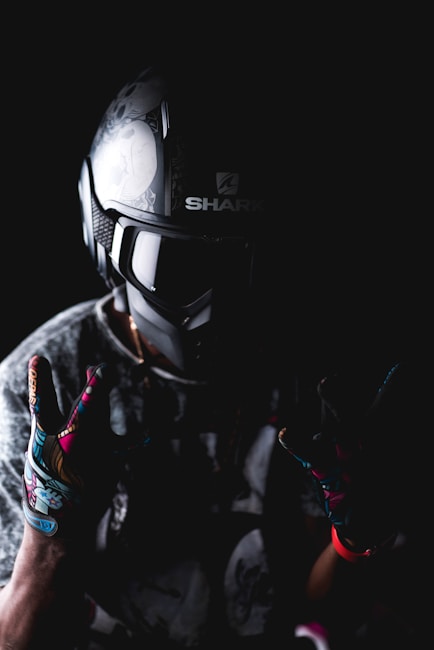A person wearing a detailed motorcycle helmet and colorful gloves is set against a dark background. The helmet has intricate designs, and the gloves feature vibrant patterns. The lighting creates a dramatic, moody effect.