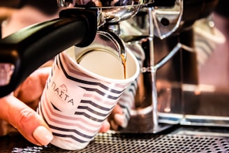 A hand holds a cup beneath a coffee machine as freshly brewed coffee pours into it. The cup is decorated with stripes and some text in a foreign script. The coffee machine is metallic, reflecting light and suggesting a modern design.