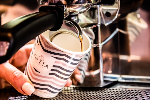 A hand holds a cup beneath a coffee machine as freshly brewed coffee pours into it. The cup is decorated with stripes and some text in a foreign script. The coffee machine is metallic, reflecting light and suggesting a modern design.