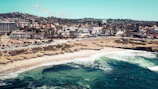 A coastal town with a sandy beach and rocky formations. Waves crash along the shoreline, where people can be seen enjoying the sun and water. In the background, there is a bustling town with numerous buildings and palm trees lining the streets.