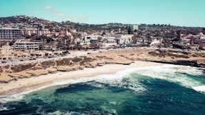 A coastal town with a sandy beach and rocky formations. Waves crash along the shoreline, where people can be seen enjoying the sun and water. In the background, there is a bustling town with numerous buildings and palm trees lining the streets.